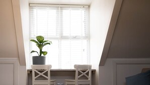 A small, modern room with a sloped ceiling, two white chairs, a round coffee table, a potted plant, and a window with blinds. - Surf Loft Steps From The Beach (Newquay)