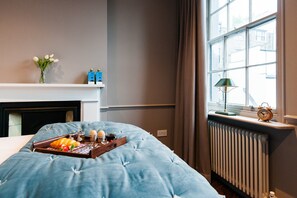 A bedroom with a bed, a bedside table with a lamp, a window with curtains, a radiator, and a fireplace. - The Cleveland Arms II (London)