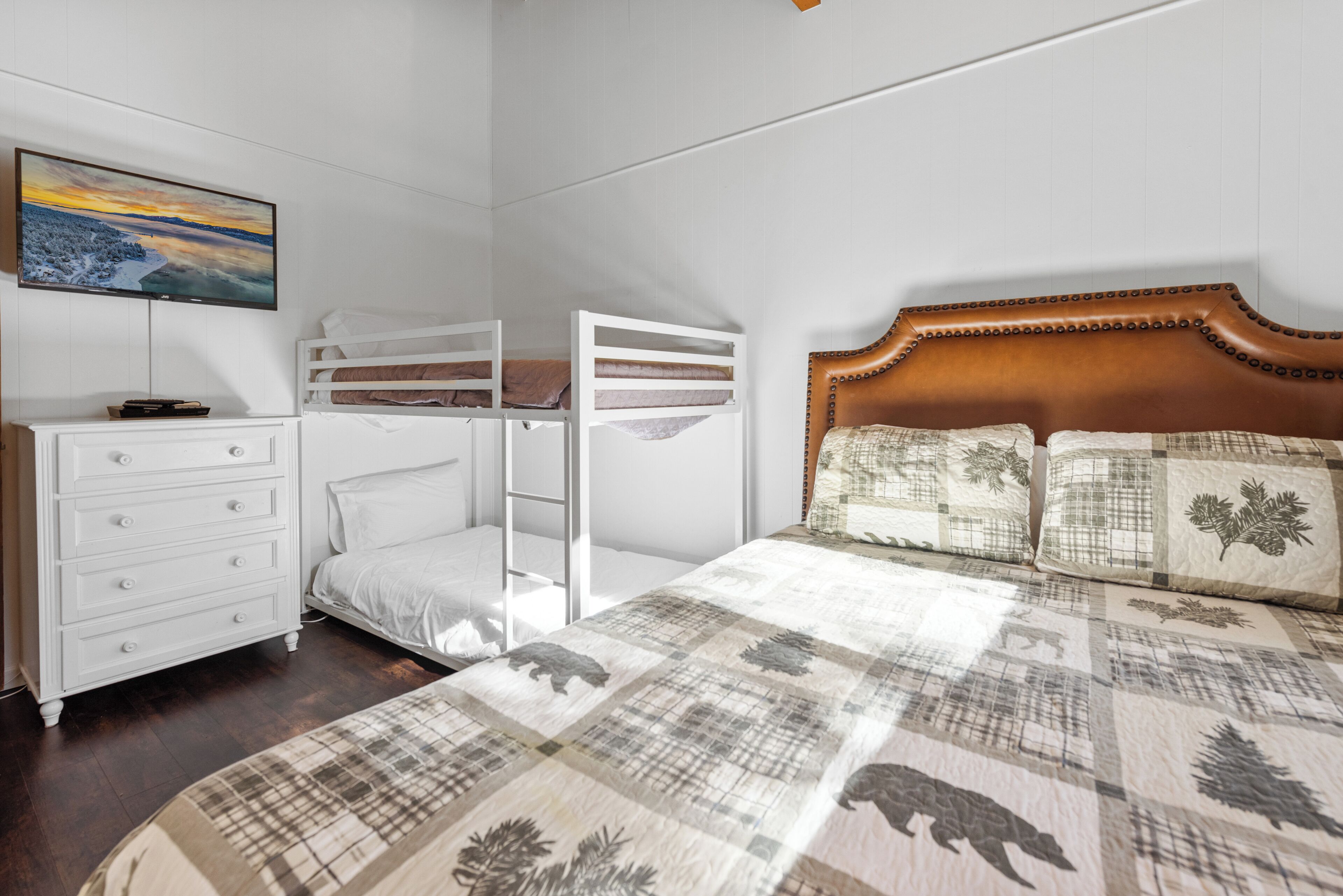 A bedroom with a bunk bed, a wooden headboard, a white dresser, and a flat-screen TV mounted on the wall.