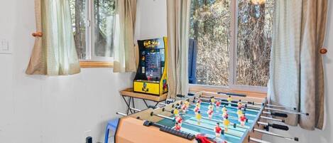 A room with a foosball table, a Pac-Man arcade machine, and a window with curtains.