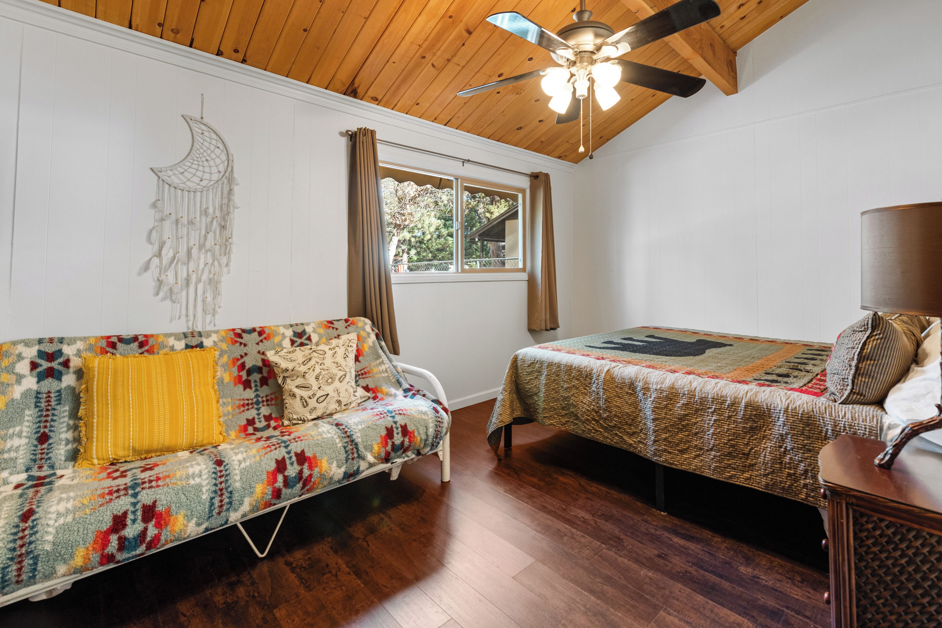 Una habitación con techo de madera, una cama con colcha estampada, un sofá con manta colorida y una ventana con cortinas.