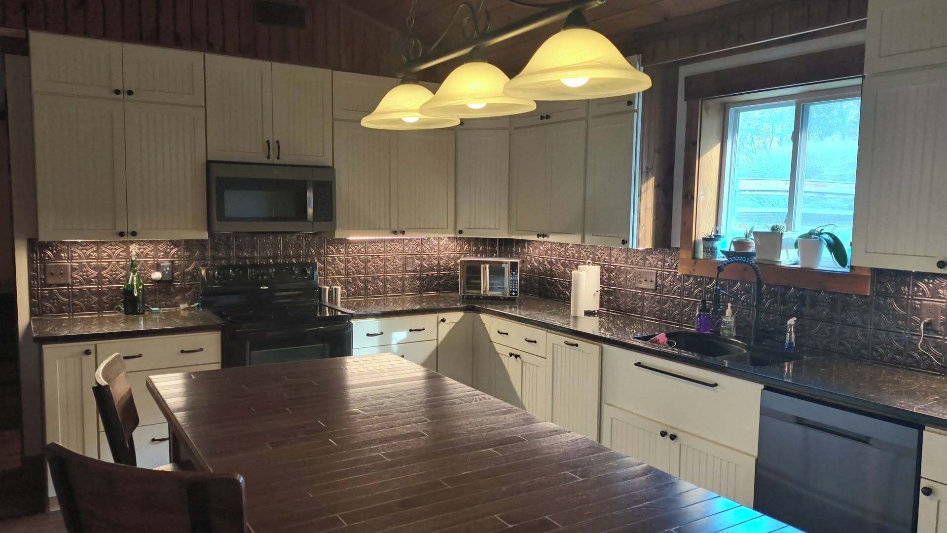 A kitchen with a wooden dining table, white cabinets, a microwave, and a window with plants.