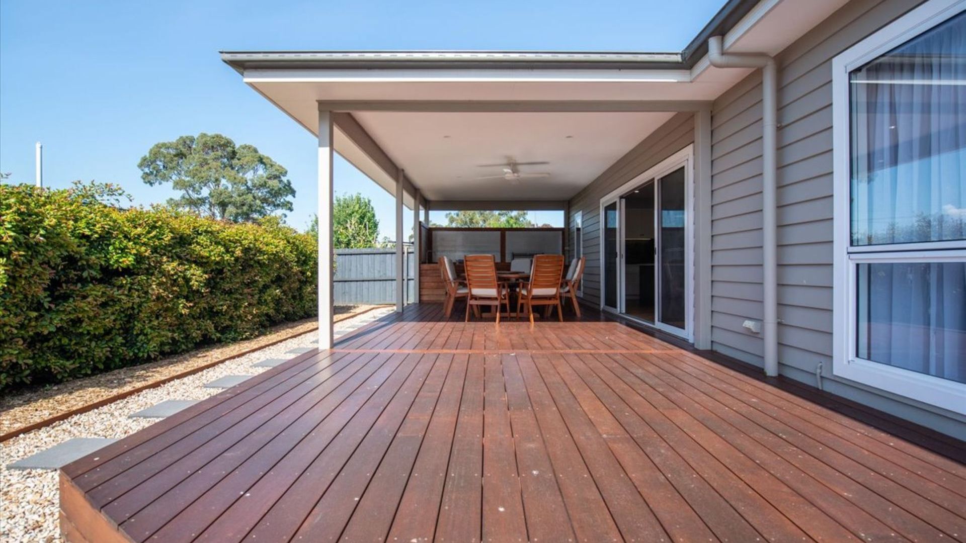 Een overdekt terras buiten, met houten vloeren, een plafondventilator en een tafel met stoelen.