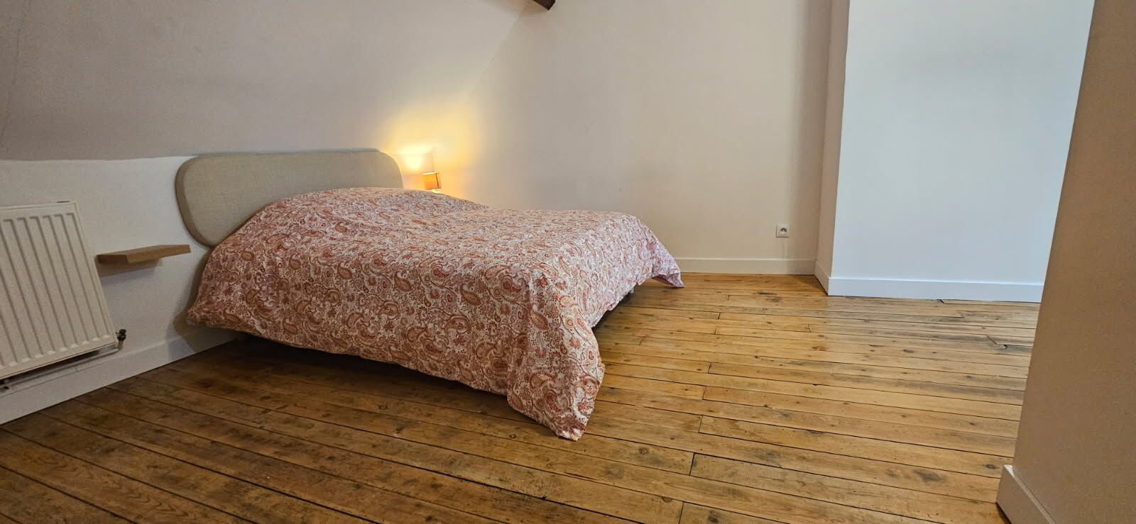 A bedroom with a wooden floor, a bed with a patterned bedspread, a bedside lamp, and a radiator.