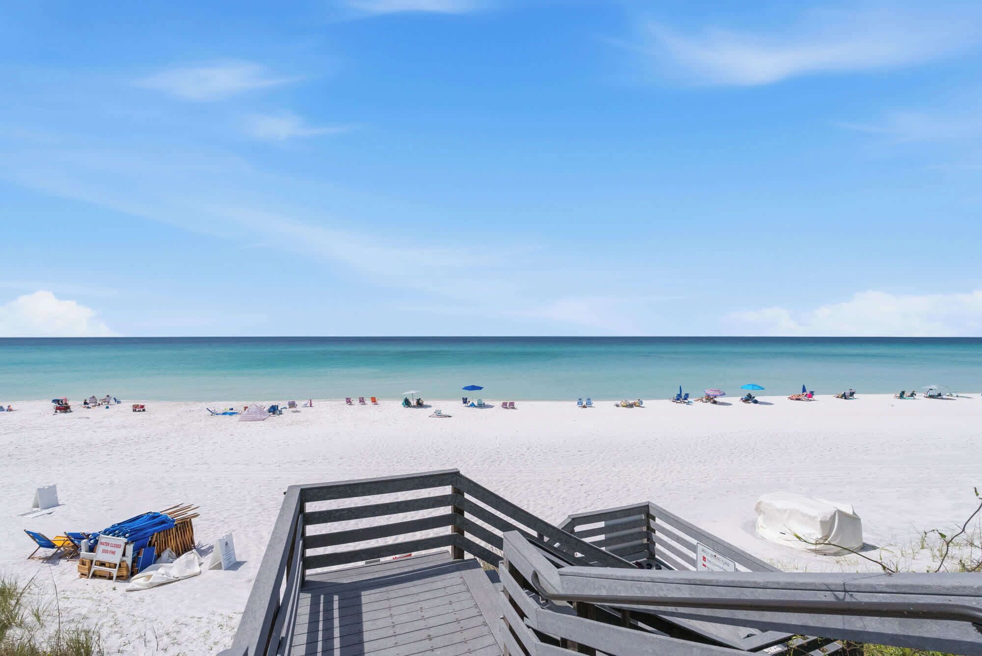 A beach with a wooden boardwalk leading to the water, clear skies, and a calm sea.