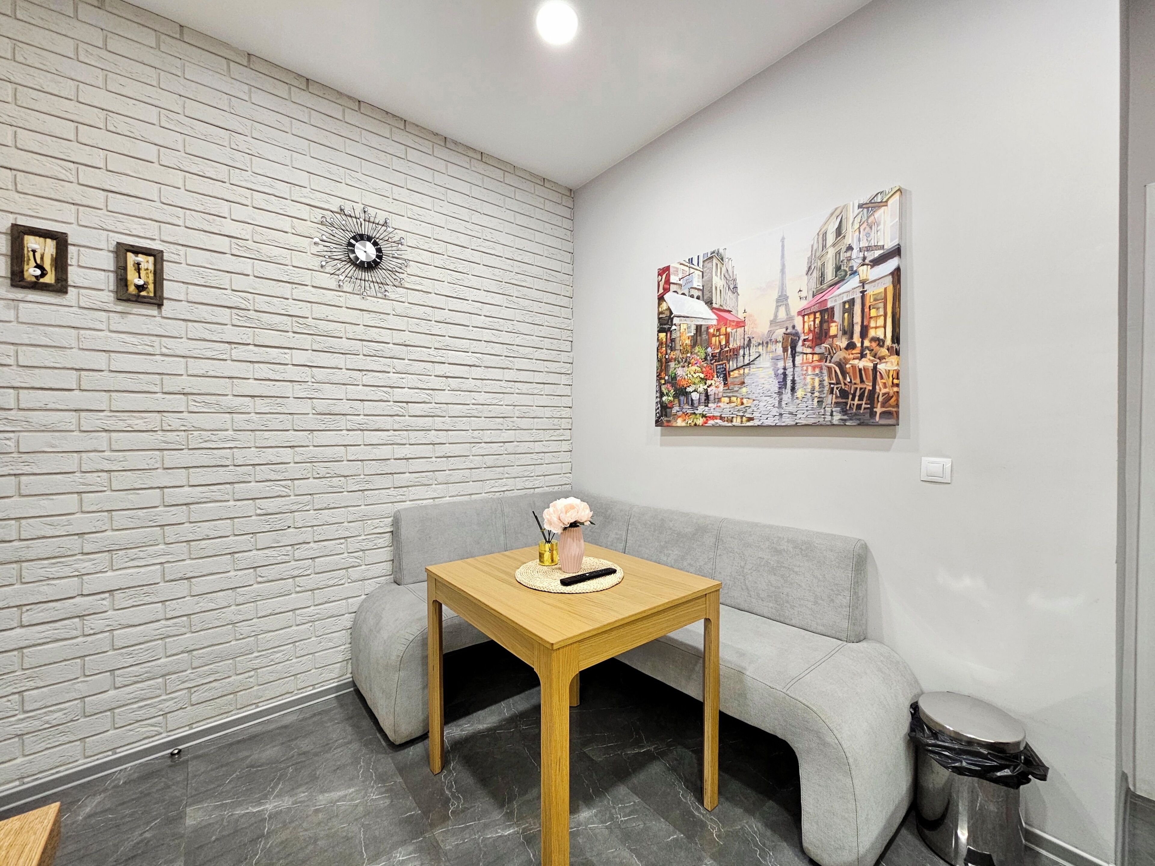 A room with a white brick wall, a grey sofa, a wooden table with a vase, a trash bin, and a painting of a street scene.