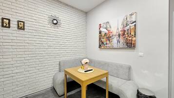 A room with a white brick wall, a grey sofa, a wooden table with a vase, a trash bin, and a painting of a street scene.