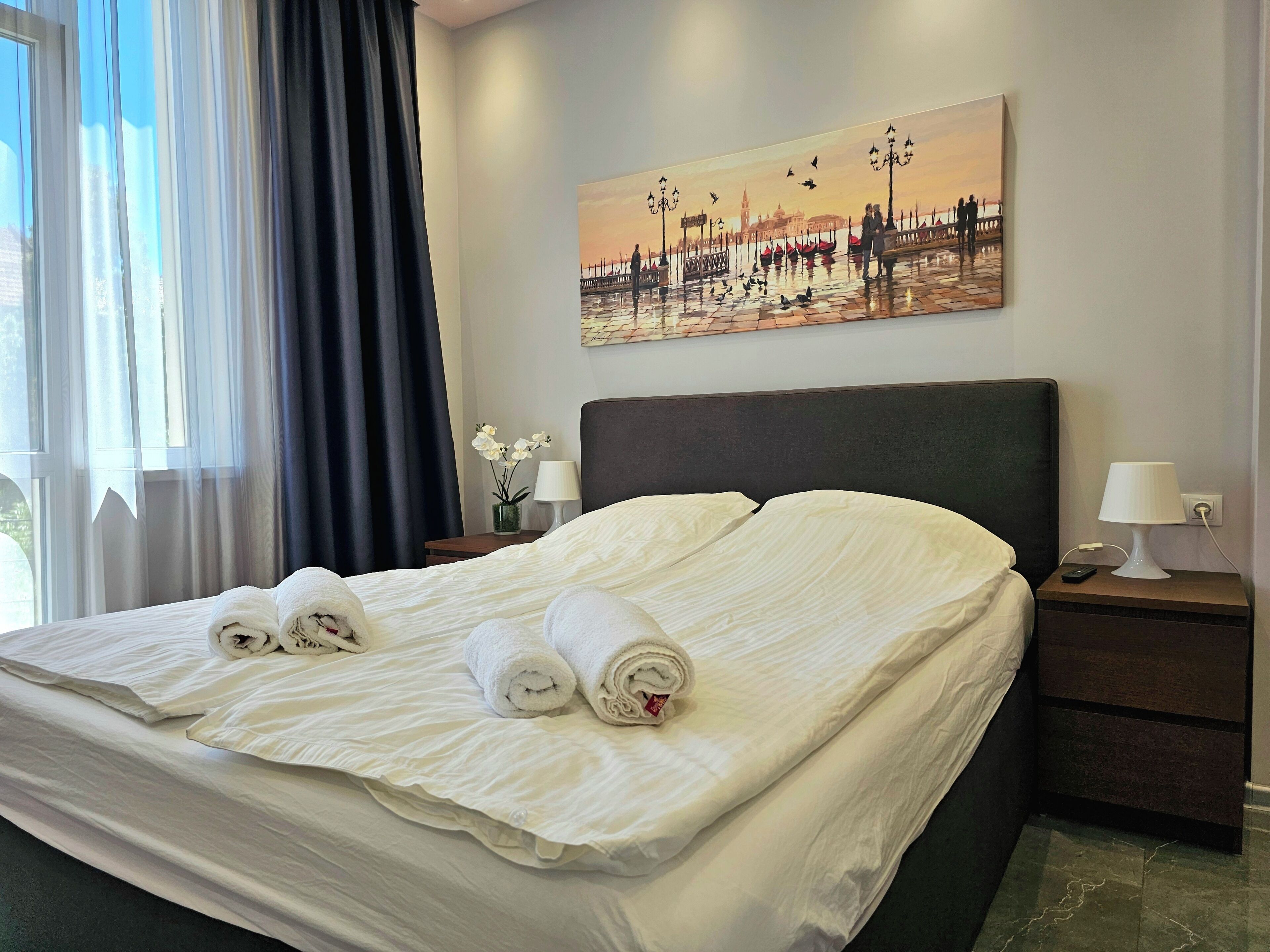 Een hotelkamer met een bed, een schilderij van een zonsondergang, een nachtkastje met een lamp en een raam met gordijnen.
