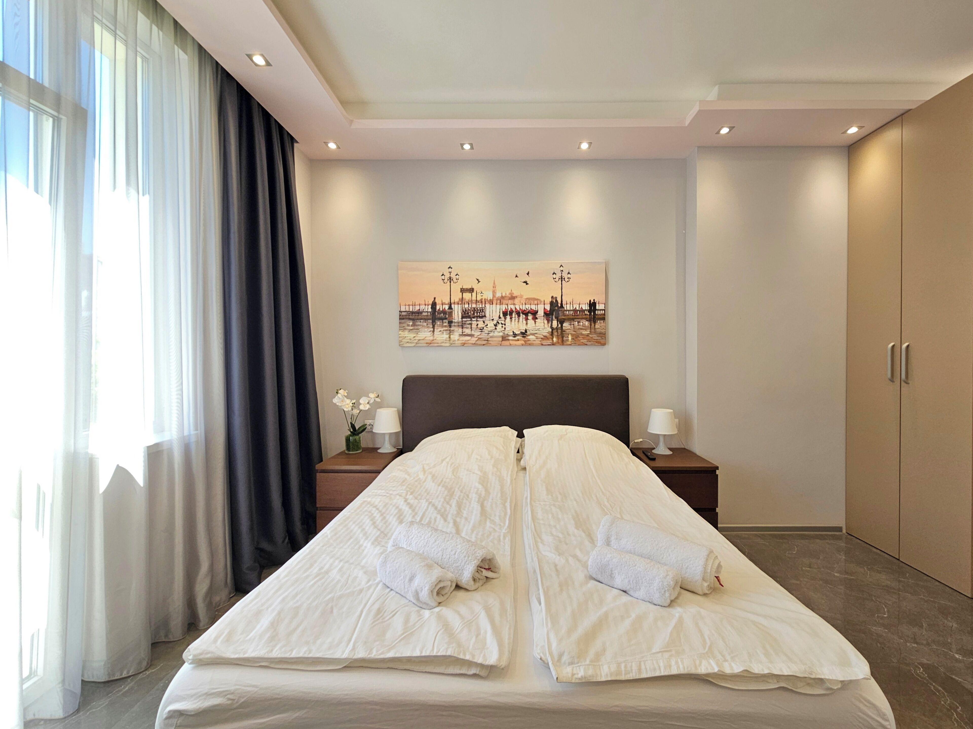 A hotel room with a large bed, a painting on the wall, a nightstand with a lamp, and a wardrobe.