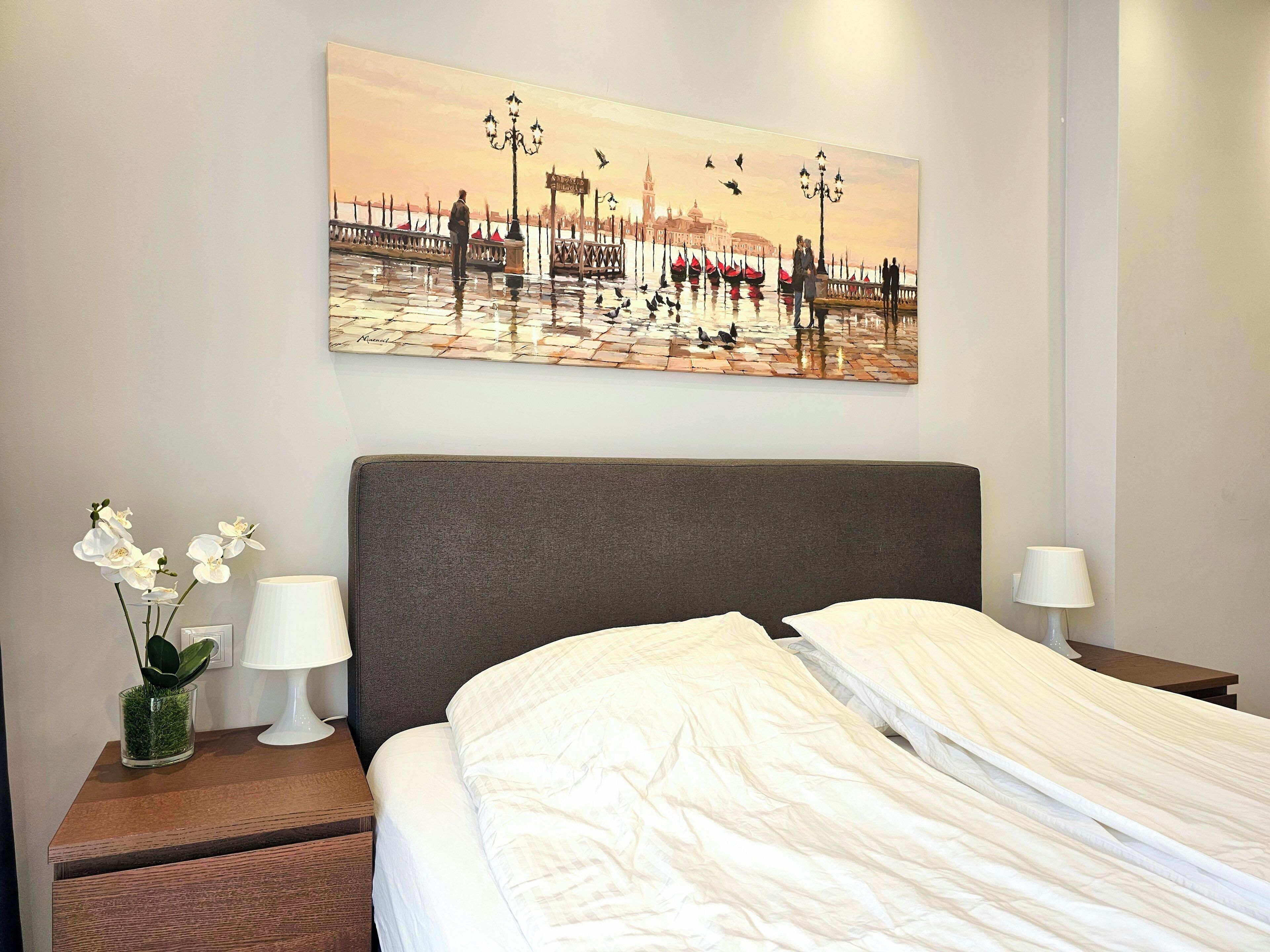 Una camera d'albergo con un letto, comodini, un vaso con fiori e un quadro che raffigura un panorama urbano.