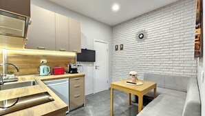 A modern kitchen with a built-in oven, a coffee machine, and a toaster. There is a small table with a lamp and a vase. A grey sofa is against the wall.