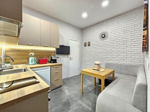 Een moderne keuken met een ingebouwde oven, een koffieapparaat en een broodrooster. Er staat een klein tafeltje met een lamp en een vaas. Een grijze bank staat tegen de muur.