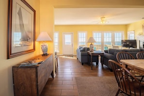 A living room with a sofa, a dining table, and a TV. There are two lamps on the table and a framed picture on the wall. - On Porpoise- Oceanfront, Pet Friendly (North Topsail Beach)