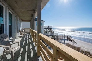 A wooden deck with chairs, a staircase leading to a beach, and a building with large windows. - On Porpoise- Oceanfront, Pet Friendly (North Topsail Beach)