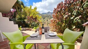 Mesa lista para dos con una botella y copas, rodeada de sillas verdes, con vistas a montañas y follaje.