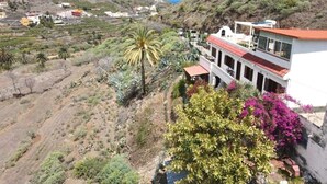 Una propiedad en la ladera de una colina con un edificio blanco que cuenta con balcones y un tejado rojo, rodeada de vegetación y un paisaje montañoso.