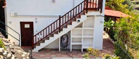 Un edificio blanco con una escalera que lleva a un balcón, rodeado de vegetación y colinas.