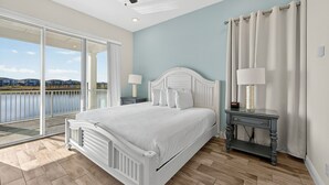 A bedroom with a large bed, a nightstand, a lamp, and a view of a body of water through a sliding glass door. - Waterfront 3BR Pool Cottage at Margaritaville Orlando - Near Disney (Kissimmee)