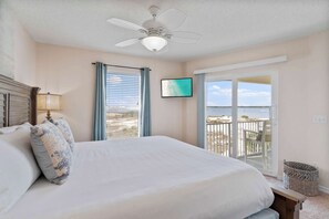 Ein Schlafzimmer mit einem Bett, einem Deckenventilator, einem Fernseher, einem Balkon mit Blick auf den Strand und einem Korbwäscheschrank.