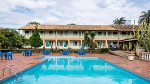 Ein Swimmingpool mit blauen Liegestühlen und Sonnenschirmen, ein Gebäude mit einem Ziegeldach und Palmen.