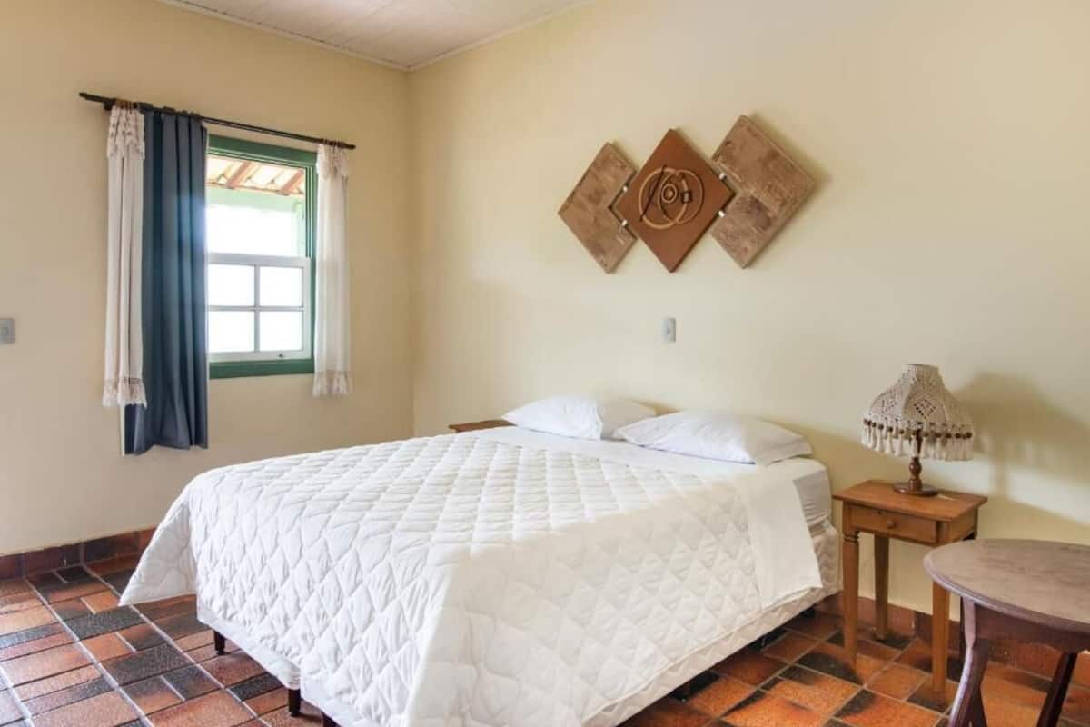 A room with a bed, a small table, a lamp, and a window with curtains.