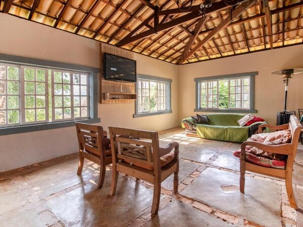 A spacious room with a high ceiling, large windows, a green sofa, and wooden chairs.
