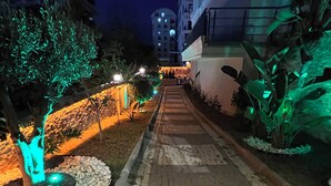 Un sendero nocturno bordeado de árboles e iluminado por luces verdes, que conduce a un edificio con varias ventanas.