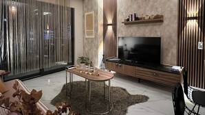 Una sala de estar moderna con televisor, muebles de madera, una mesa de comedor y un ventanal con cortinas.