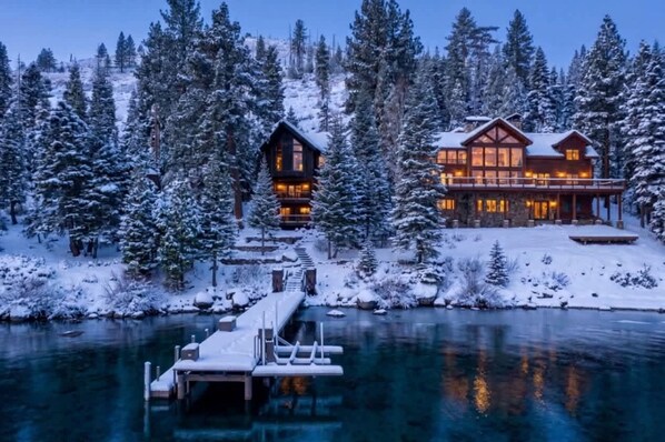 A large lodge with multiple levels, surrounded by snow-covered trees and a dock extending into a calm body of water.