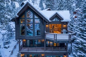 A multi-story wooden cabin with large windows, a balcony, and a deck, surrounded by snow-covered trees.