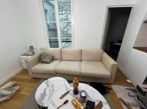A living room with a beige sofa, a round marble coffee table, and a window with a view of a tree. - Modern Apartment 1br/4p - Paris 10th (Paris)