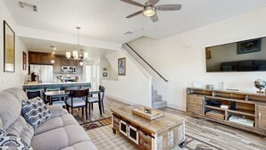 A modern living room with a sofa, coffee table, and TV. There's a kitchen area with a dining table and chairs. A staircase leads to another level.