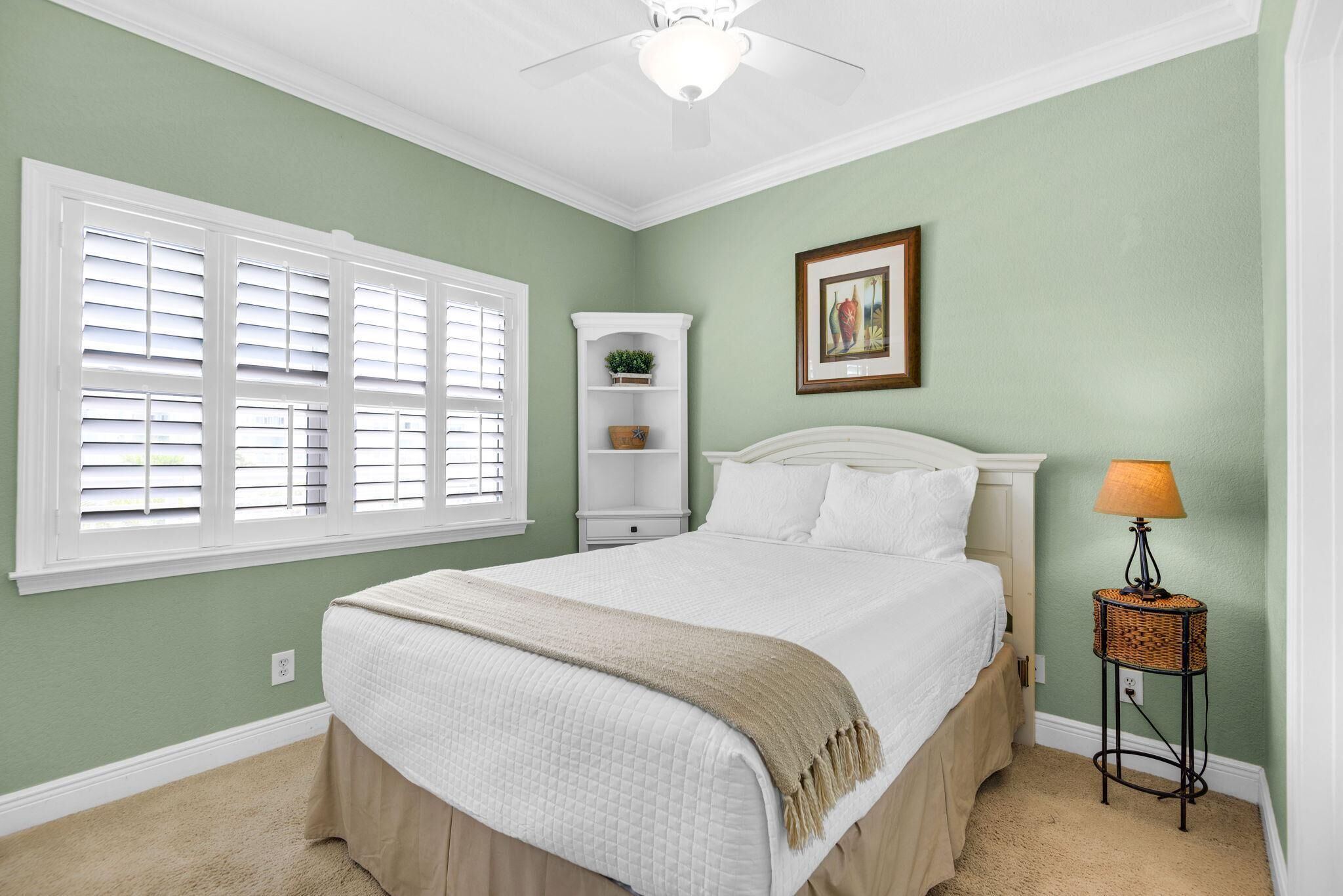 A bedroom with a bed, a window with blinds, a bedside lamp, a shelf with a plant, and a framed picture on the wall.
