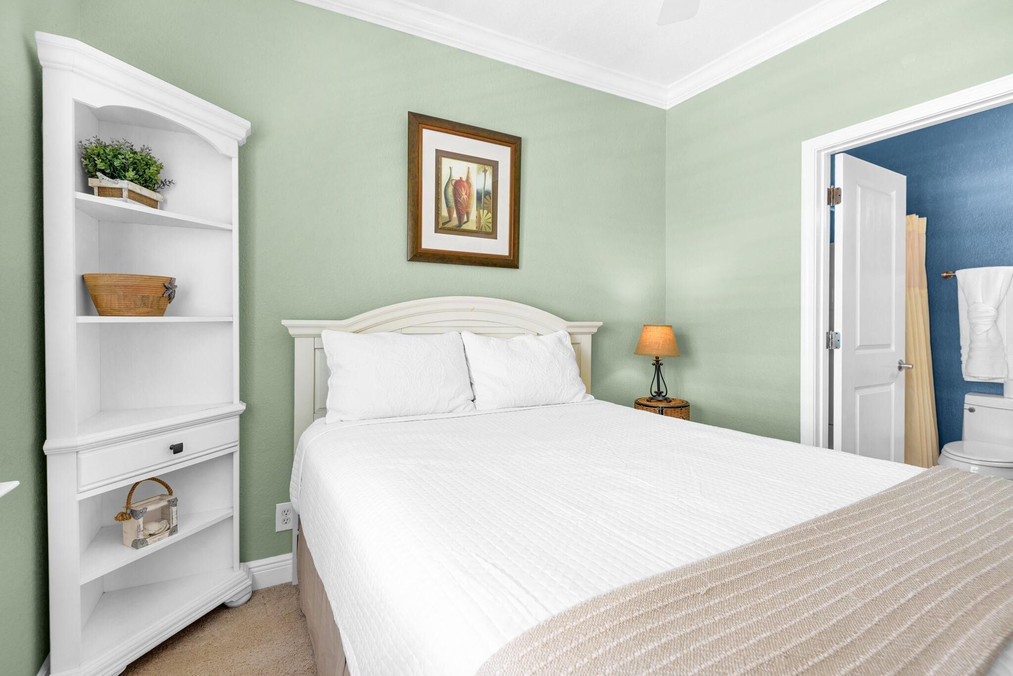 A bedroom with a bed, a nightstand, a shelf unit, a framed picture, and a door leading to a bathroom.