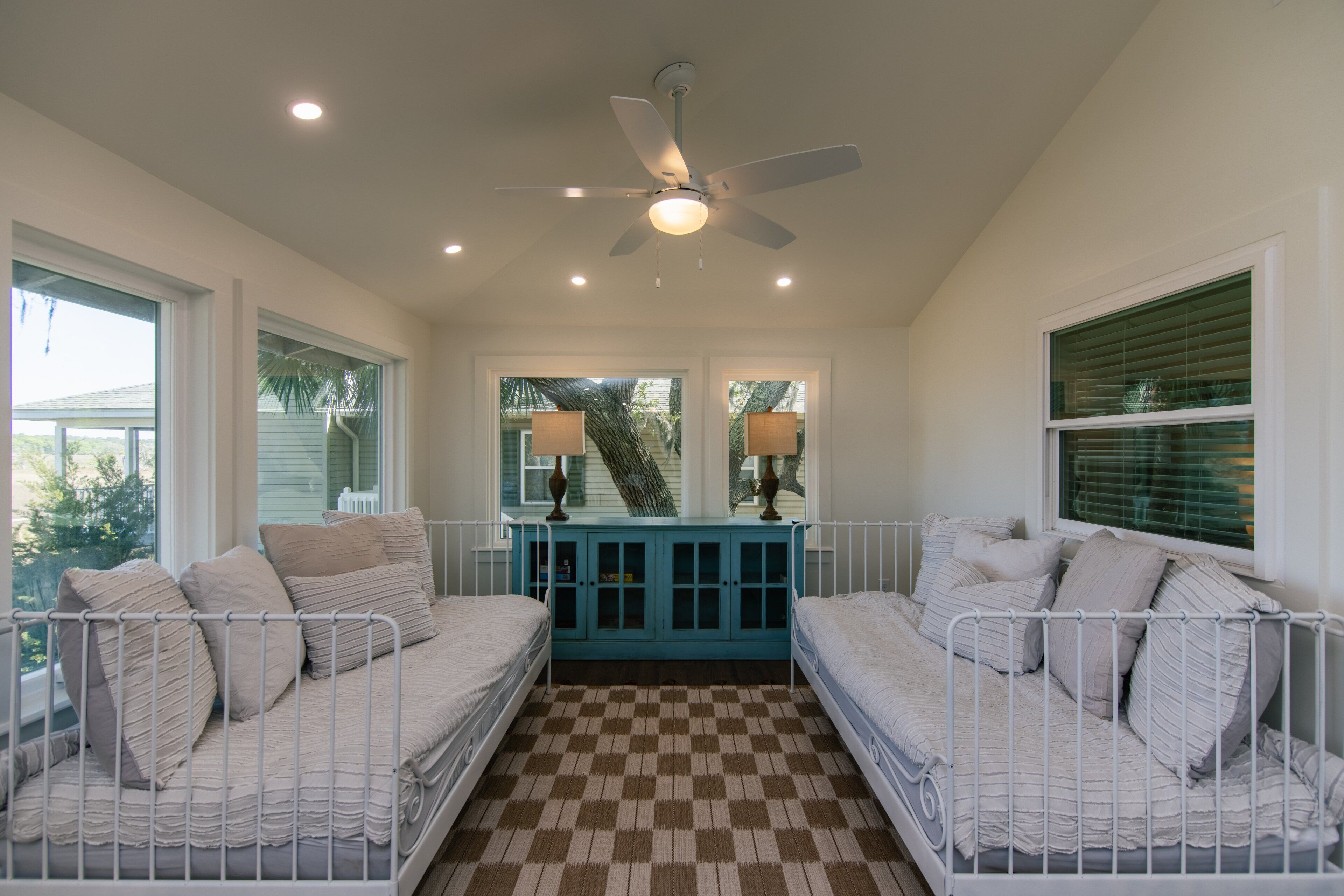 A room with two beds, a ceiling fan, a window with blinds, and a checkered floor.