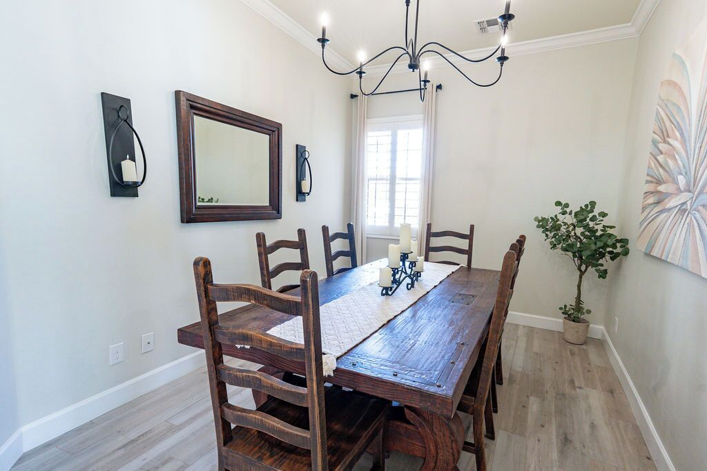 A dining room with a wooden table, six chairs, a chandelier, a wall-mounted mirror, and a potted plant.