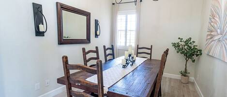 A dining room with a wooden table, six chairs, a chandelier, a wall-mounted mirror, and a potted plant.