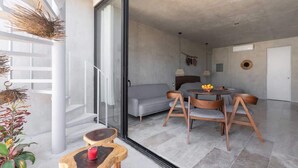 Ein modernes Interieur mit einer Glastür, die auf einen Balkon führt, ein graues Sofa, Holzstühle, ein kleiner runder Tisch und ein Blumentopf.