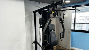 A gym with a bench, weight machine, and a window with a view of buildings. - Elegance Suítes Ibirapuera Moema (São Paulo)