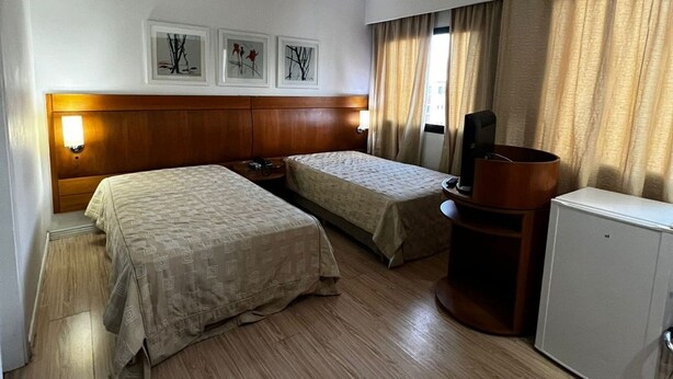 A hotel room with two beds, a wooden headboard, a small desk, a television, and a refrigerator. - Elegance Suítes Ibirapuera Moema (São Paulo)