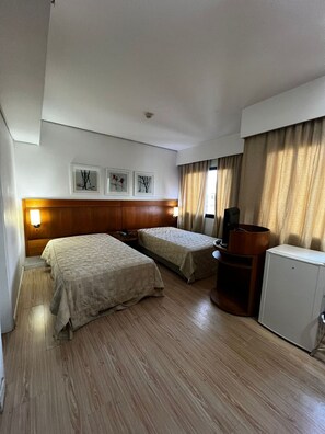 A hotel room with two beds, a wooden headboard, a small desk, a television, and a refrigerator. - Elegance Suítes Ibirapuera Moema (São Paulo)