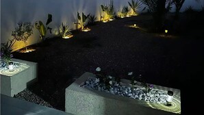 Een verlichte buitenruimte 's nachts, met een witte muur, palmbomen en verlichte plantenbakken.