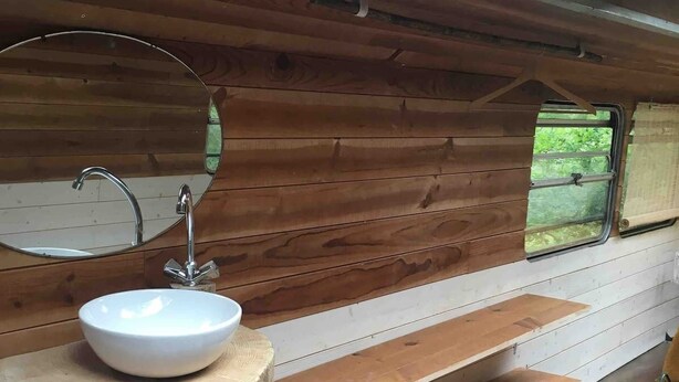 Interior de cabana de madeira com pia redonda de madeira, um espelho, uma janela com vista e um banquinho de fibra natural.