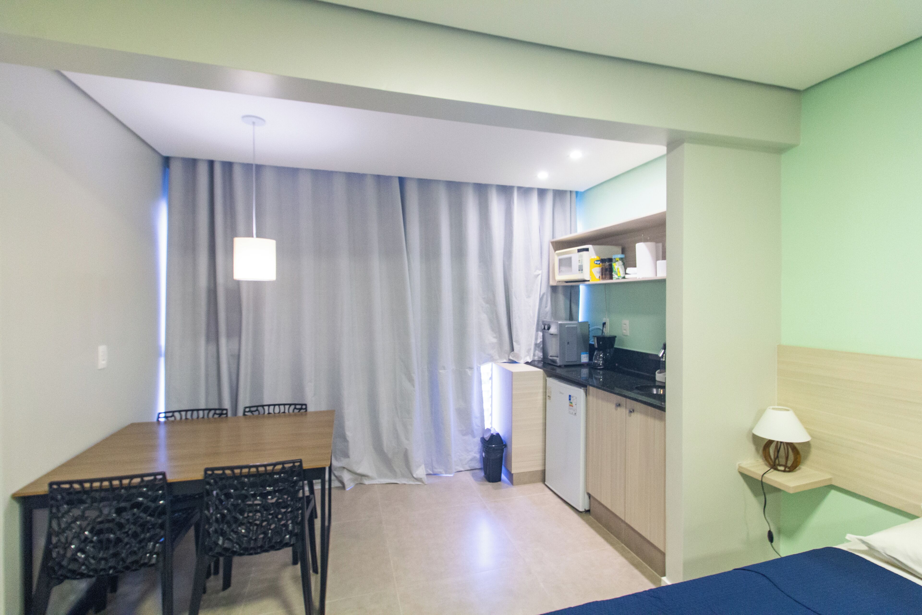 Habitación compacta con mesa de comedor, sillas, cama, kitchenette pequeña y ventana con cortinas.