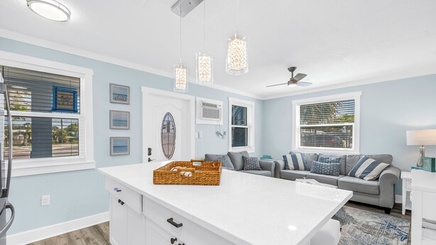 Une cuisine moderne avec des meubles blancs, un îlot central, des luminaires suspendus et un canapé gris dans le salon attenant.