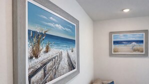 Una stanza con un quadro grande raffigurante una spiaggia, un quadro più piccolo e un divano beige con cuscini blu.