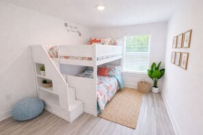 A child's bedroom with bunk beds, a small staircase, a window with blinds, a plant, and framed pictures on the wall.