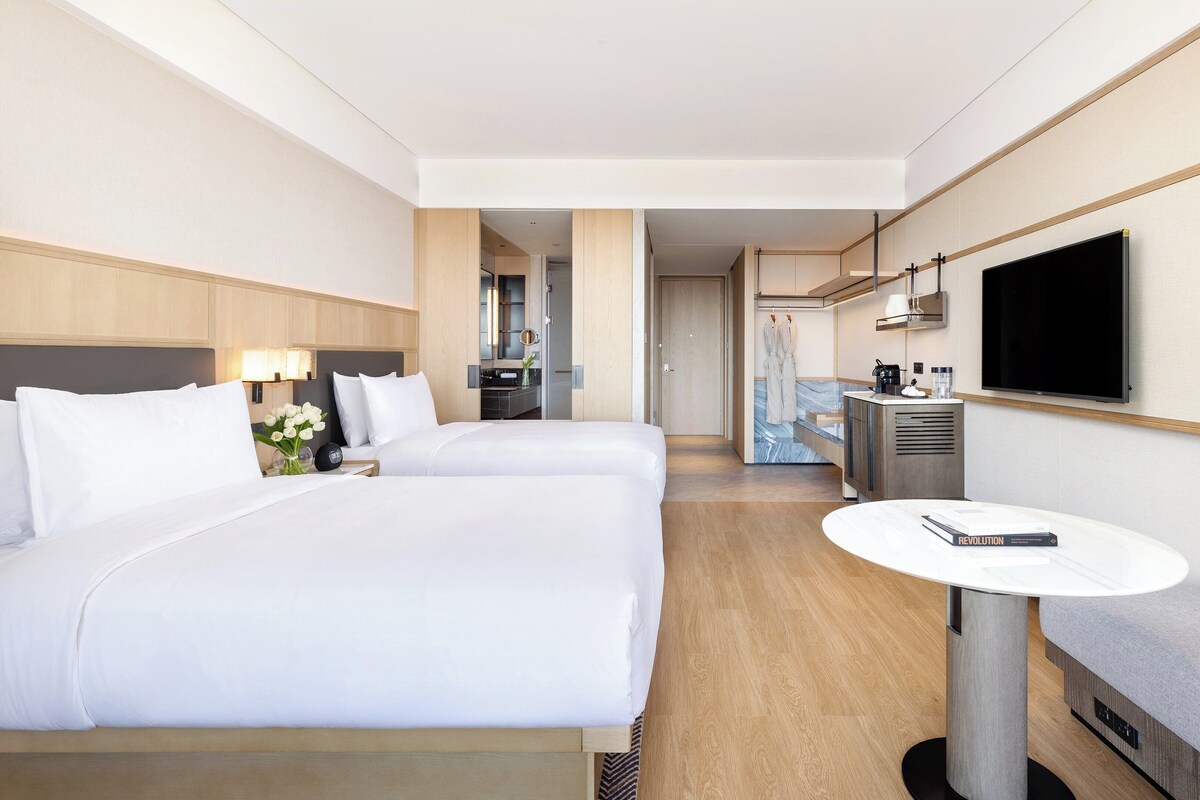 Une chambre d’hôtel avec deux lits, une télévision, une petite table, un canapé et une salle de bain visible à travers une porte ouverte.