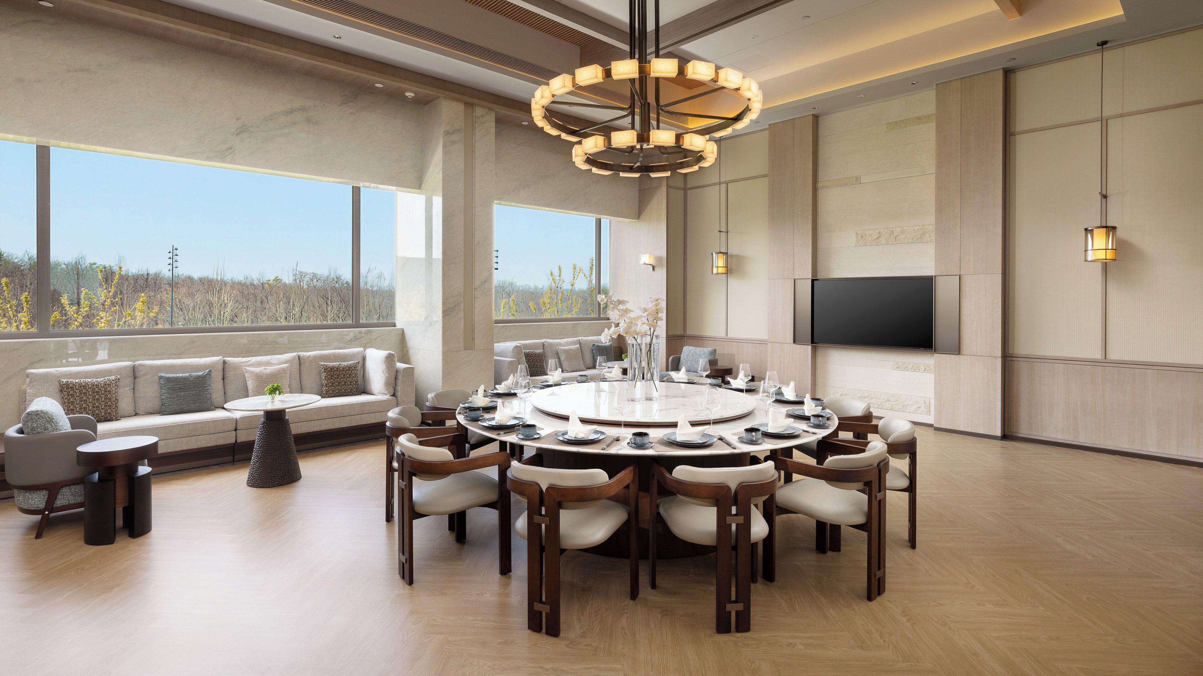 A modern dining room with a round table, comfortable seating, a large window with a view, and a flat-screen TV mounted on the wall.