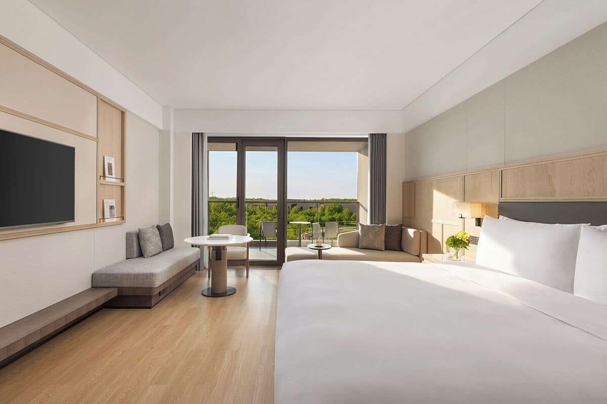 A modern hotel room with a large bed, a sofa, a small table, and a TV. There is a balcony with a view of the outdoors.